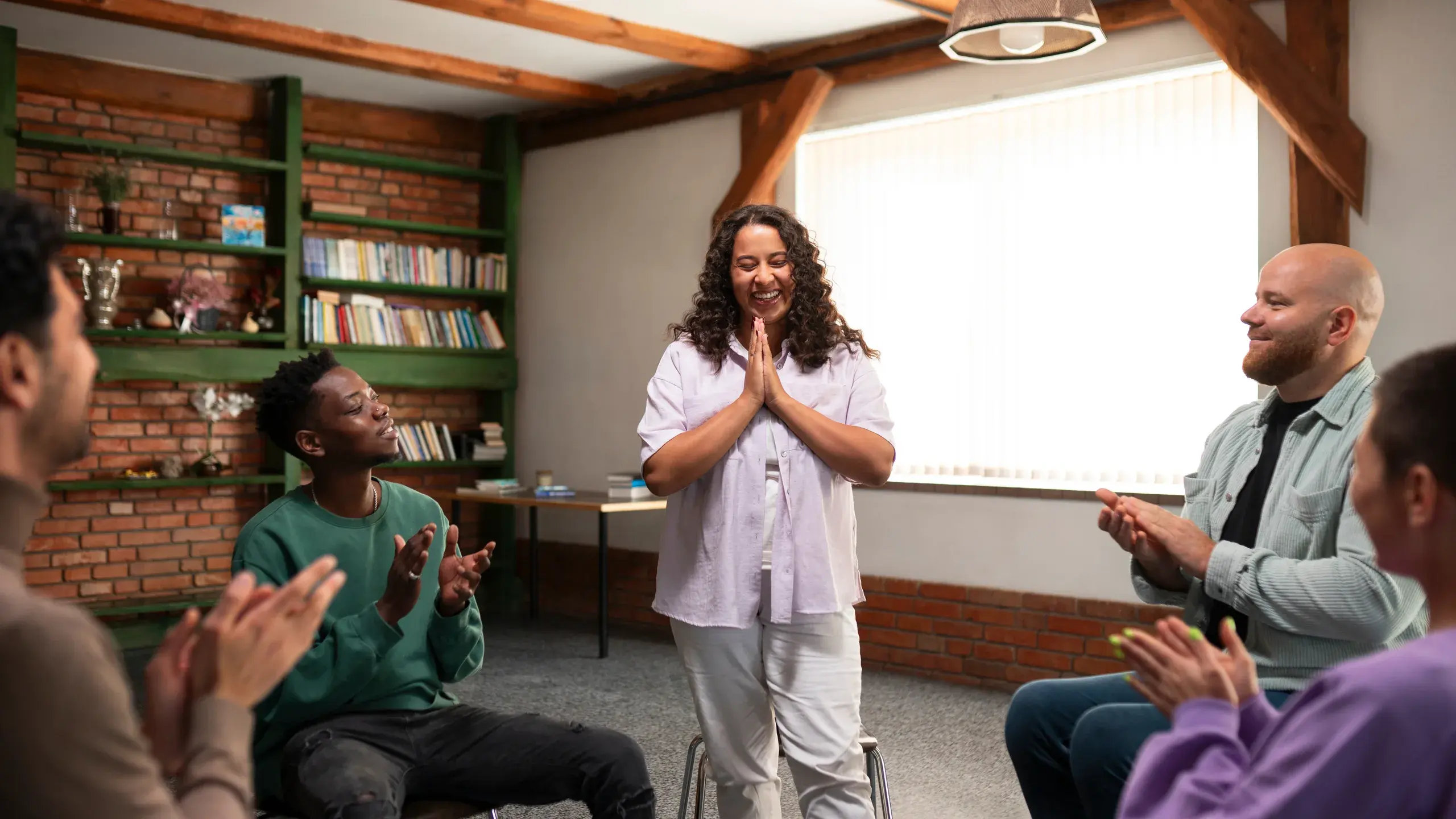 A small church group in a library room, smiling and clapping as one woman shares, hands pressed together